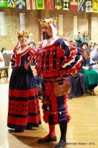 Baronessa Maria Pagani and Baron Juan Xavier tribtue to the Patriots was a team effort.  It was a team effort. Pani Astrida Borowska made Maria's dresst. Lady Syele von Heidelberg made Juan's Wams and coif. Juan made the pants. Elvira Violante de Ballesteros made the red/white/blue cording. The coif slashes represent the team logo and the large “X” motif on the back of the Wams is both a play on my name, Xavier, and also makes him #10 on the team. Maria’s gown has elements drawn from a Patriots cheerleader uniform.